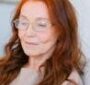 Aged female with red hair in eyeglasses looking at screen of smartphone and making online video call on blurred background